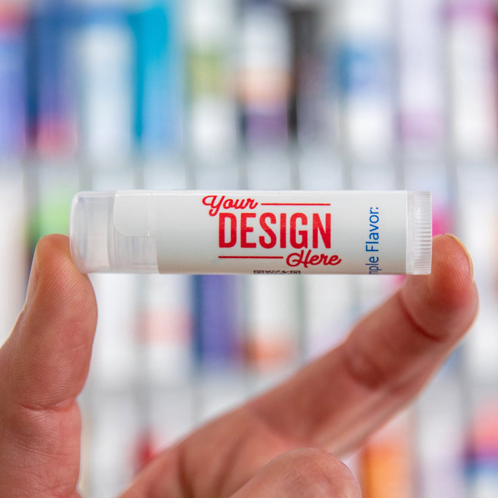 Hand holding a customizable lip balm tube with 'Your Design Here' text against a blurred background of similar lip balm tubes.
